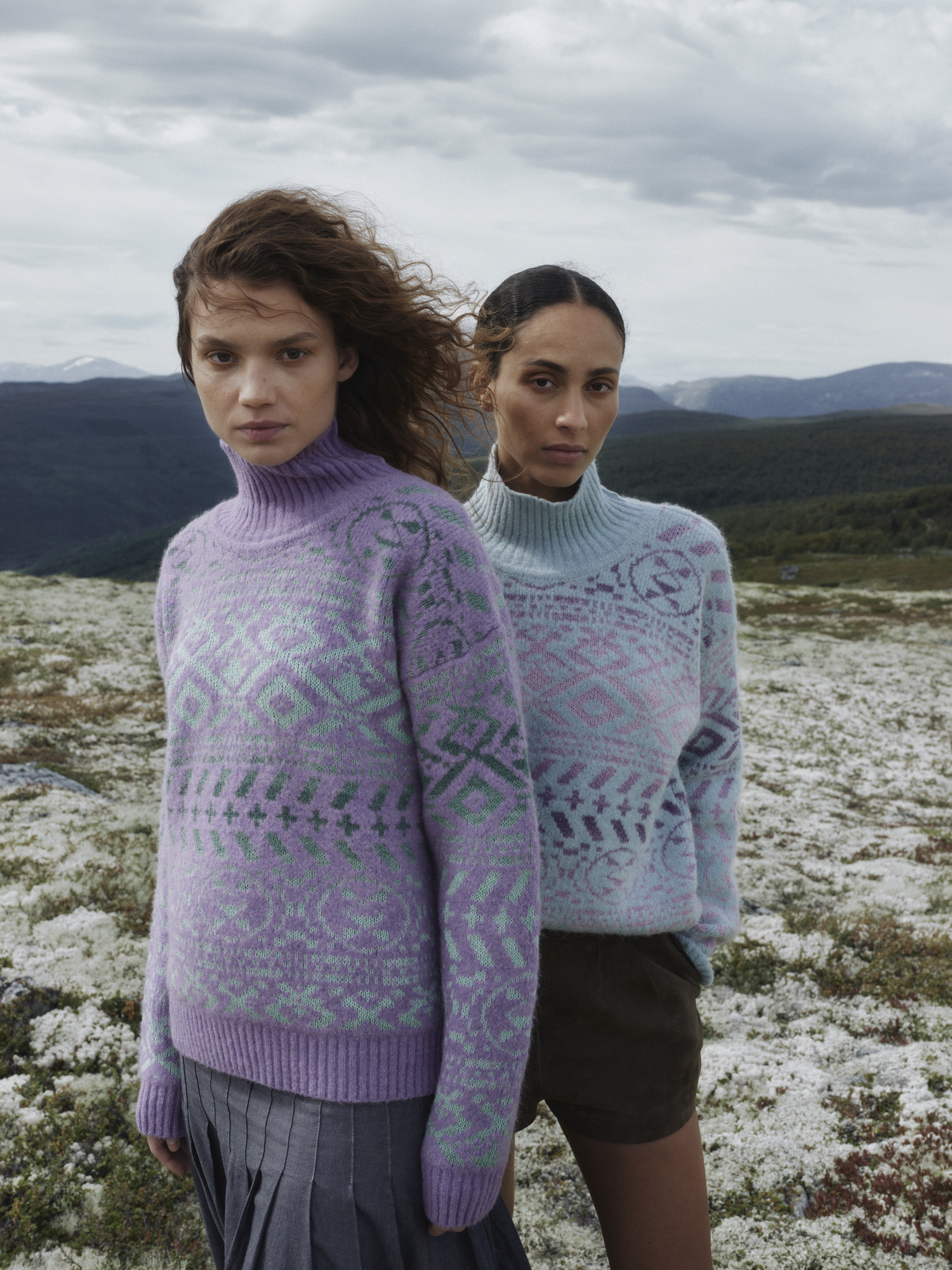 To modeller iført Highalnd ullgensere for dame fra Dale of Norway i pastellfarger, stående i fjellandskap med lyng, under dramatisk himmel