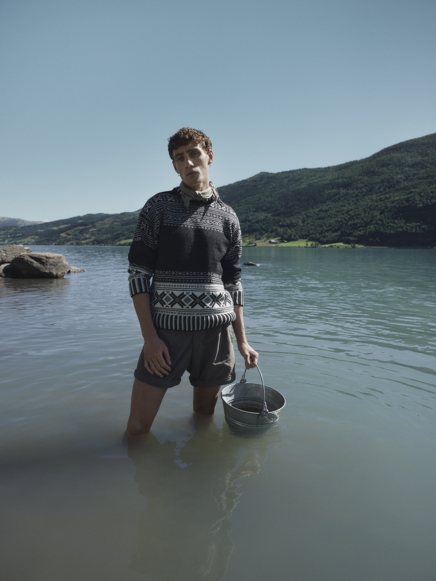 Modell i ført mørk ullgenser med tradisjonelt mønster fra Dale of Norway, stående i fjordvann med bøtte i hånda, norsk natur i bakgrunnen